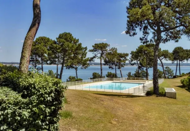 Piscine au milieu d’un jardin arboré avec une étendue d’eau visible entre les pins.