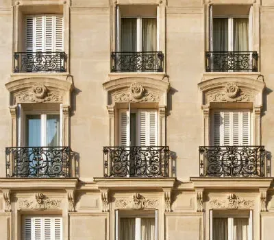 Façade ouvragée d’un immeuble ancien en pierre claire avec balcons en fer forgé et volets blancs.
